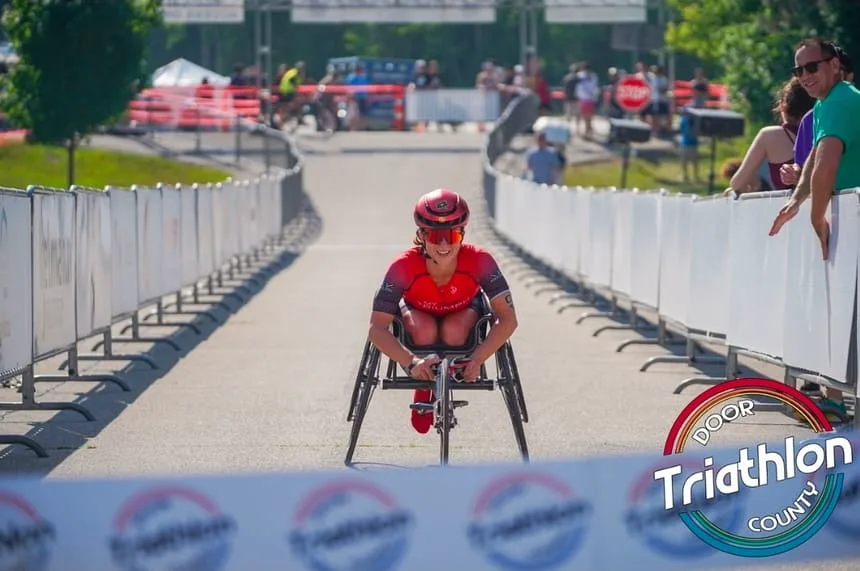 An athlete in a red racing chair driving toward the finish of the Door County Triathlon, tucked low between the barriers with spectators watching from the roadside