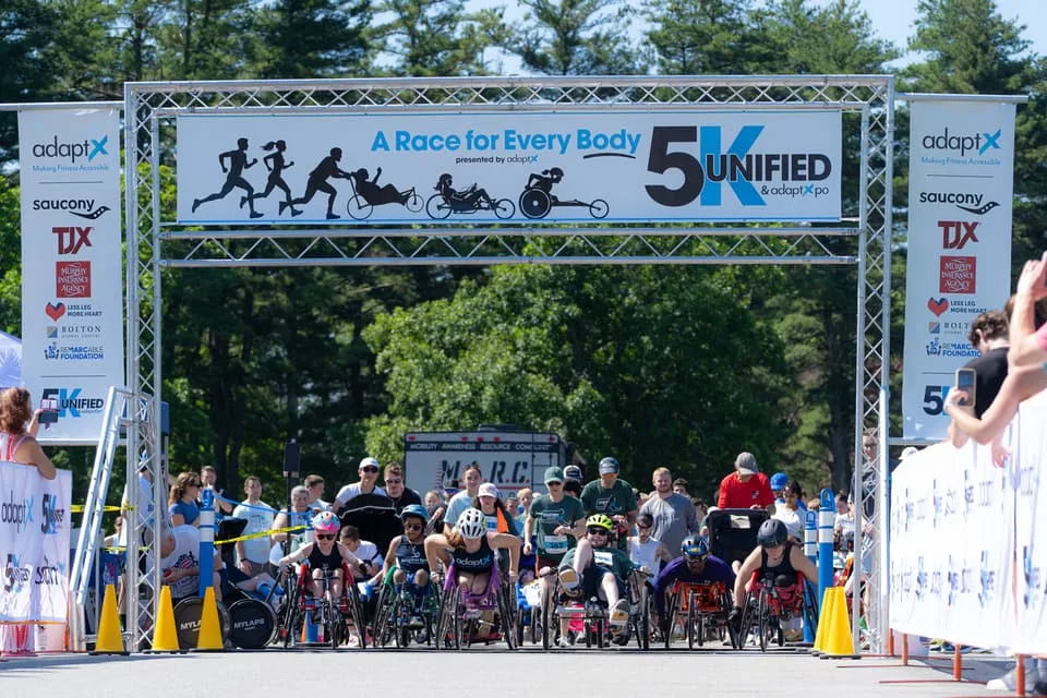 The start of the Unified 5K in Lancaster, Massachusetts: a front row of push-rim racers and handcyclists waiting under an arch reading A Race for Every Body, with a crowd of runners behind them