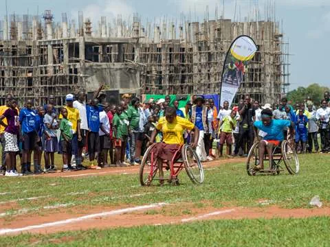 Two athletes in racing wheelchairs on a grass field, with a long line of participants waiting behind them